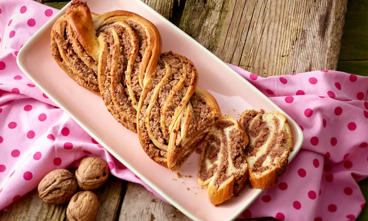 Geflochtener Nuss-Strudel mit goldbrauner Kruste und lockerer Füllung, serviert auf rosa Teller, umgeben von rosa Tuch und Walnüssen
