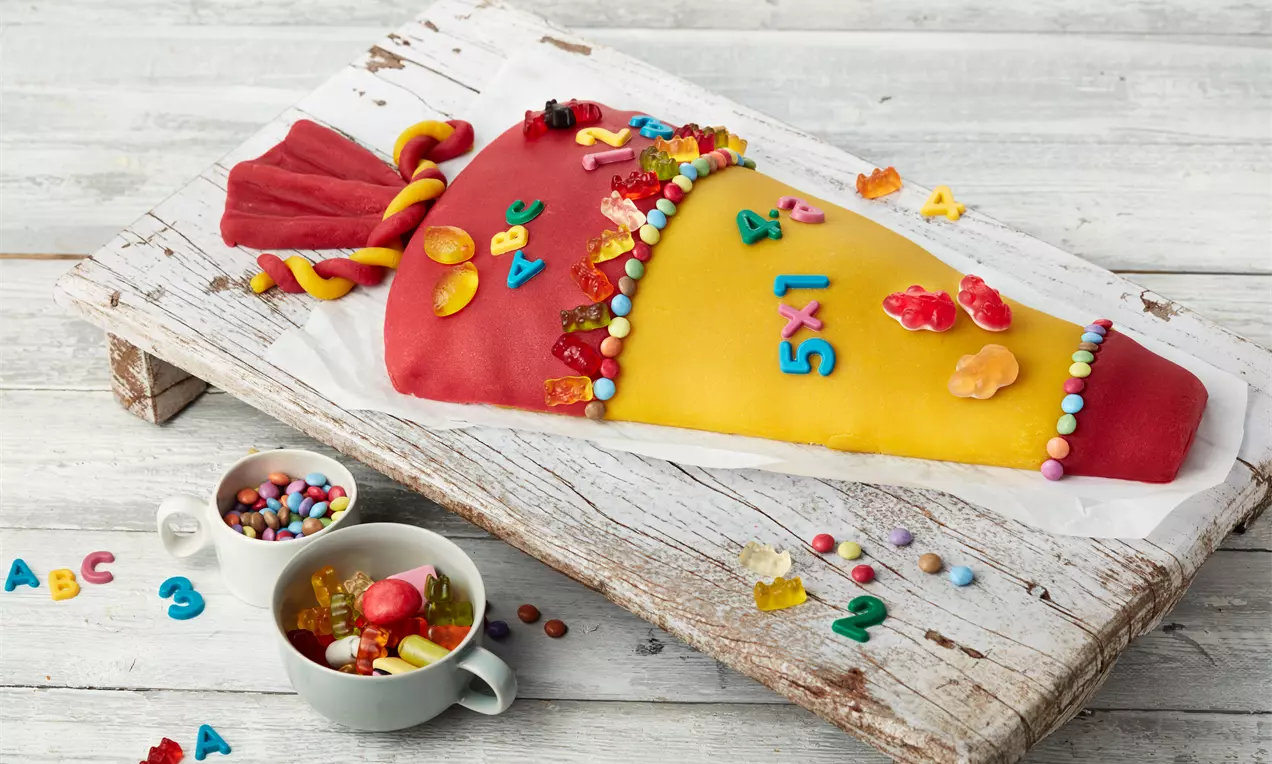 Bunte Schultüte aus roter und gelber Fondantmasse, dekoriert mit Zuckergusszahlen, bunten Perlen und Fruchtgummis auf rustikalem Holzbrett