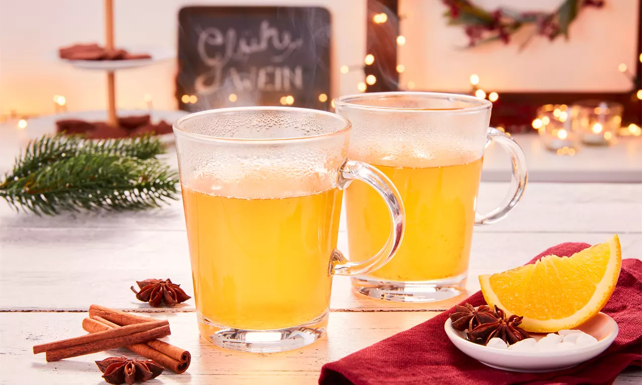 Zwei gläserne Tassen mit dampfendem, goldenem Glühwein, arrangiert mit Zimtstangen, Sternanis, Orangenscheiben und rotem Stoffserviettendekor