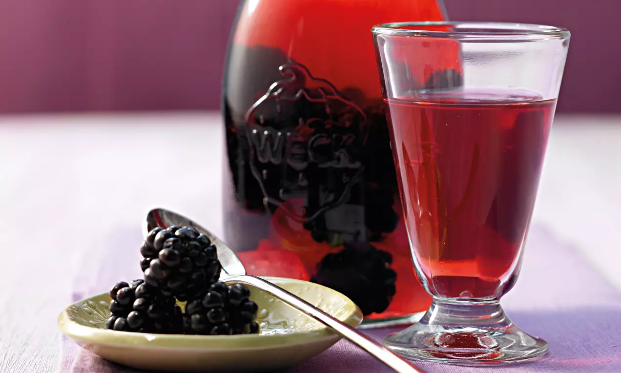 Tiefroter Brombeer-Likör im Glas neben einer bauchigen Flasche, begleitet von glänzend schwarzen Brombeeren auf einem kleinen Teller mit Silberlöffel