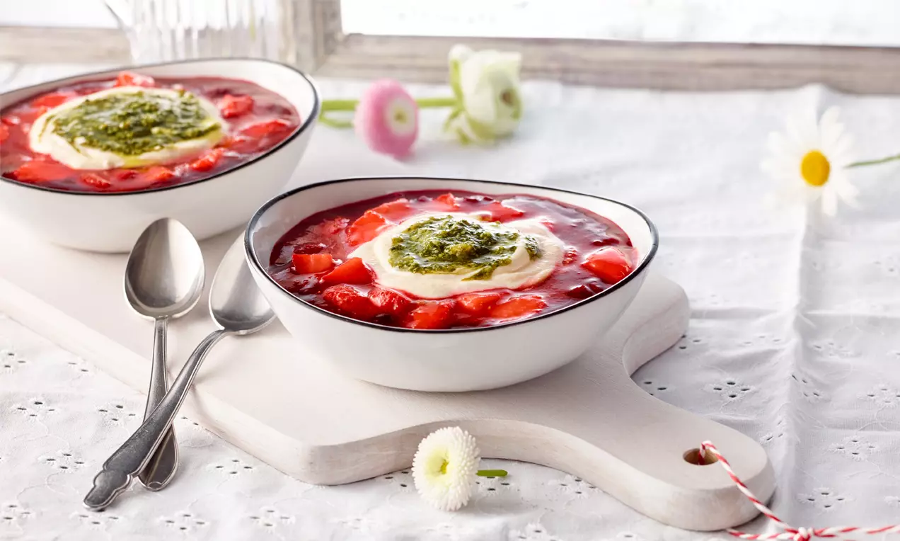 Zwei weiße Schalen mit glänzender roter Grütze, marinierten Erdbeerstücken, Sahnehaube und grünem Pesto auf hellem Holzbrett