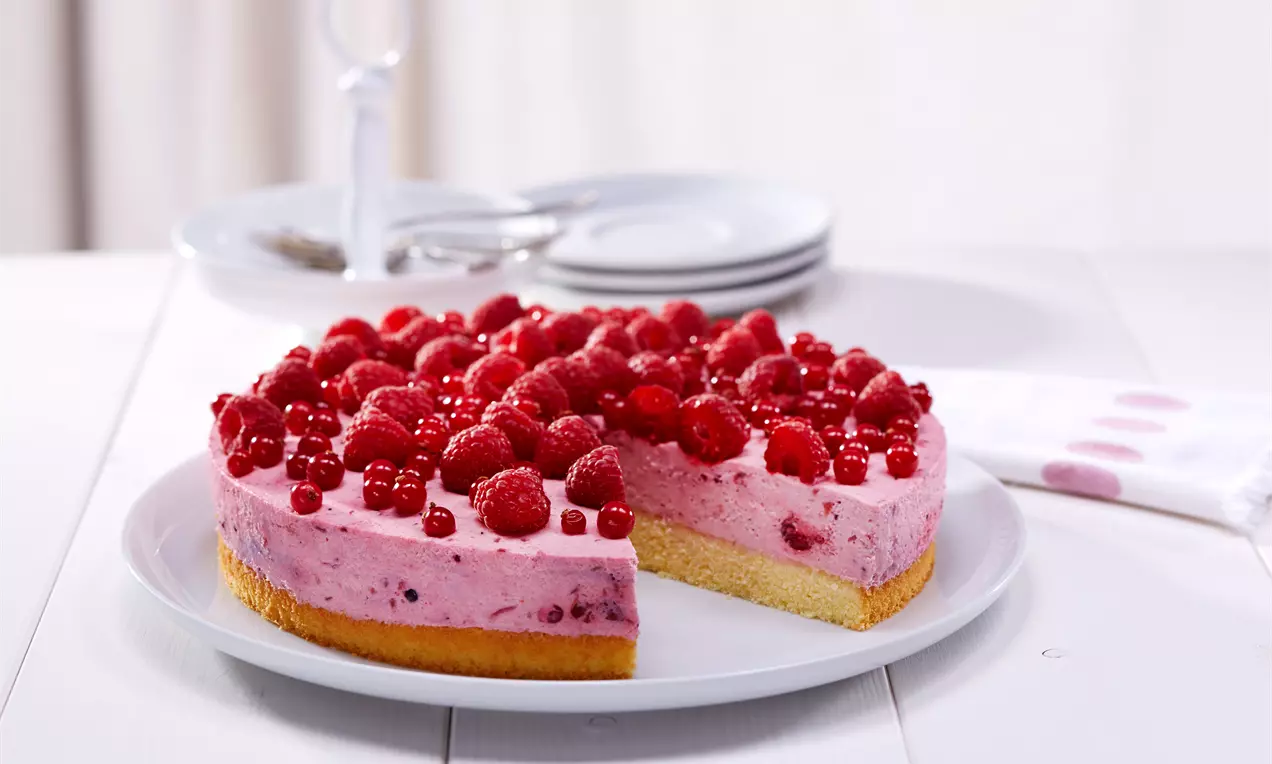 Rosa Rote-Grütze-Creme mit frischen Himbeeren und Johannisbeeren auf hellem Biskuitboden, serviert auf weißer Kuchenplatte