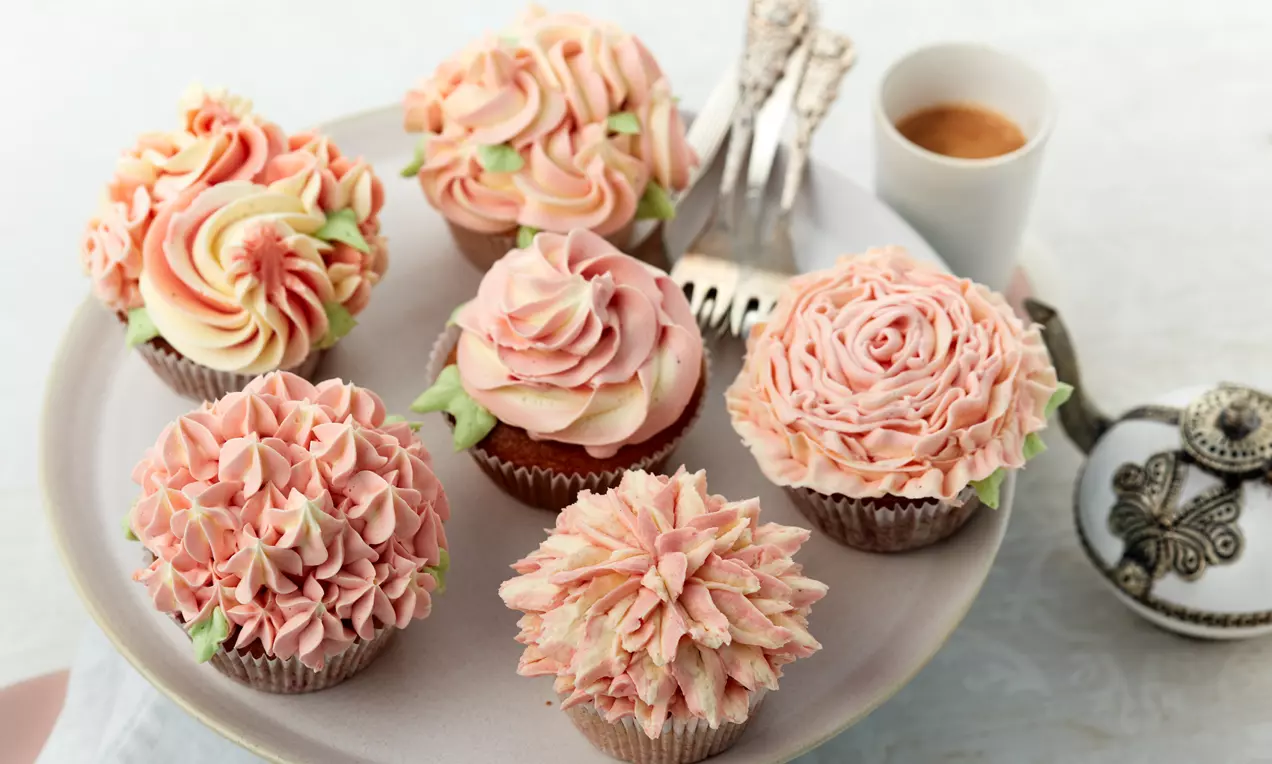 Cupcakes mit cremefarbenen und rosafarbenen, blütenförmig aufspritzen Texturen dekorativ arrangiert auf einer hellen Platte mit Besteck und Espresso