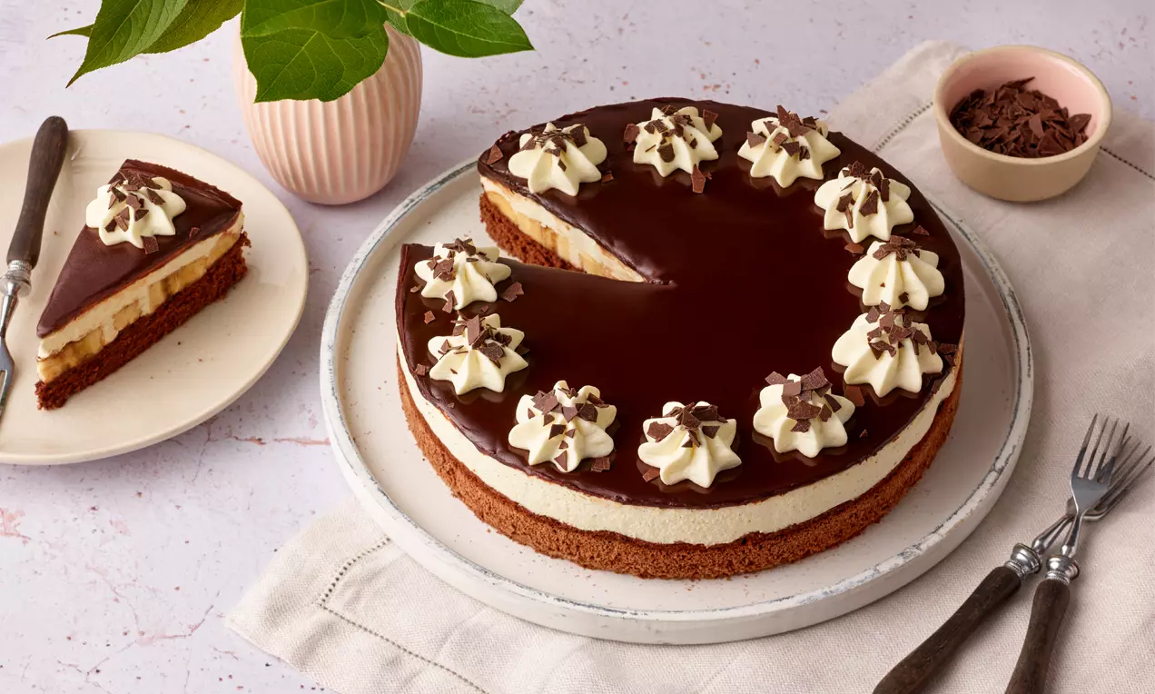 Schoko-Bananentorte mit glänzender Schokoglasur, Sahnetupfen und Schokoraspeln auf weißer Platte, daneben angeschnittenes Stück