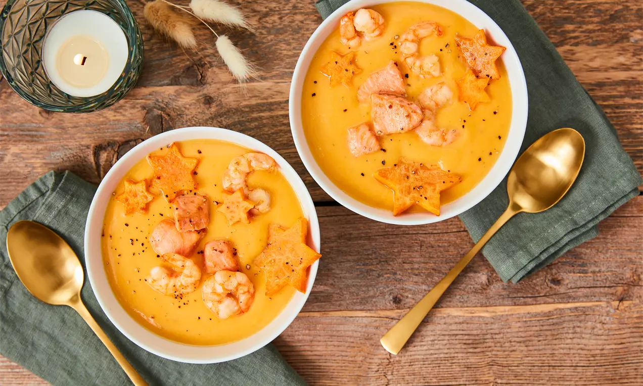 Cremige orange Süßkartoffelsuppe in weißen Schalen, garniert mit Garnelen, Lachs und sternförmigen Kroketten auf Holztisch mit grünen Servietten