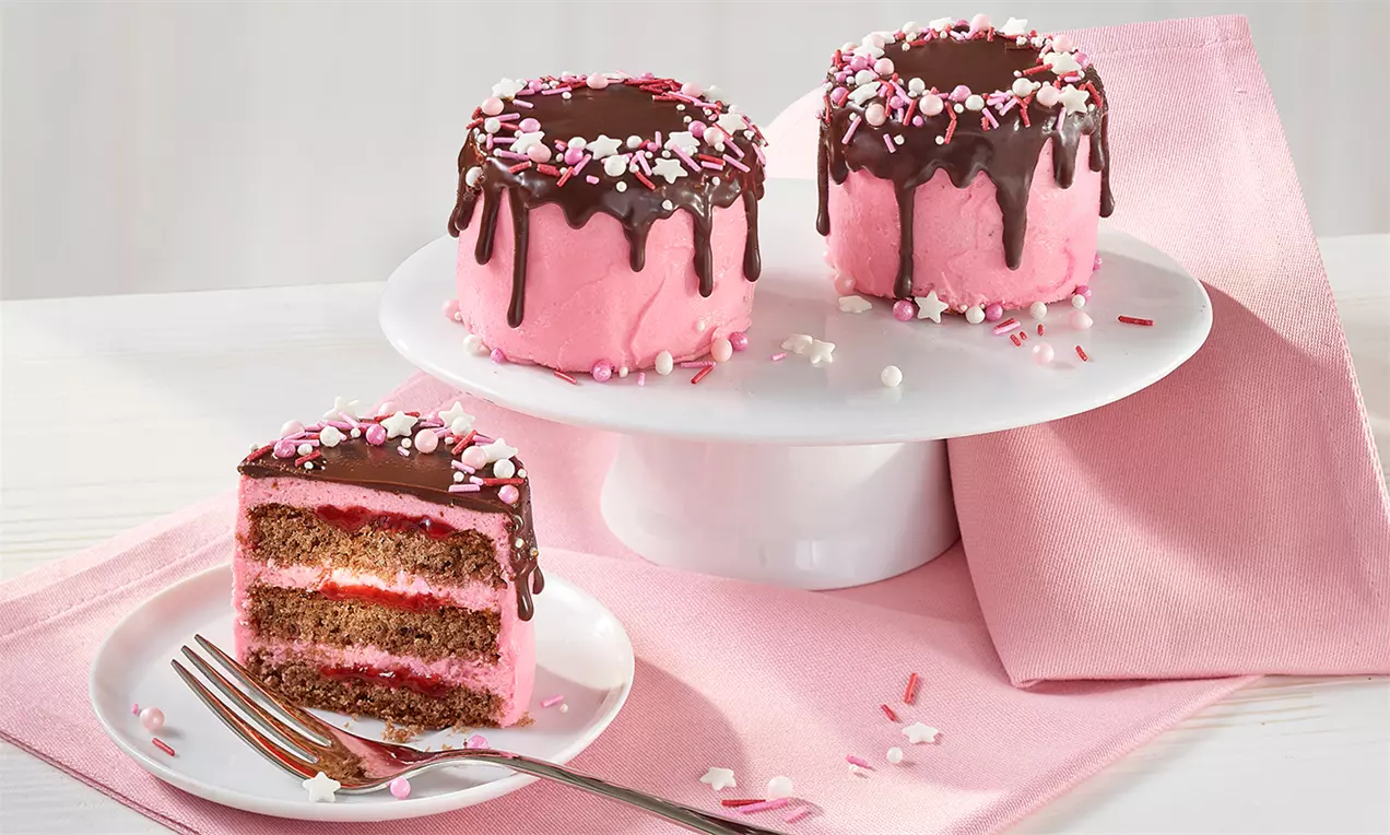 Zarte pinke Törtchen mit Schokoladenglasur und bunten Streuseln auf Tortenplatte, daneben angeschnittener Kuchen auf Teller mit Gabel