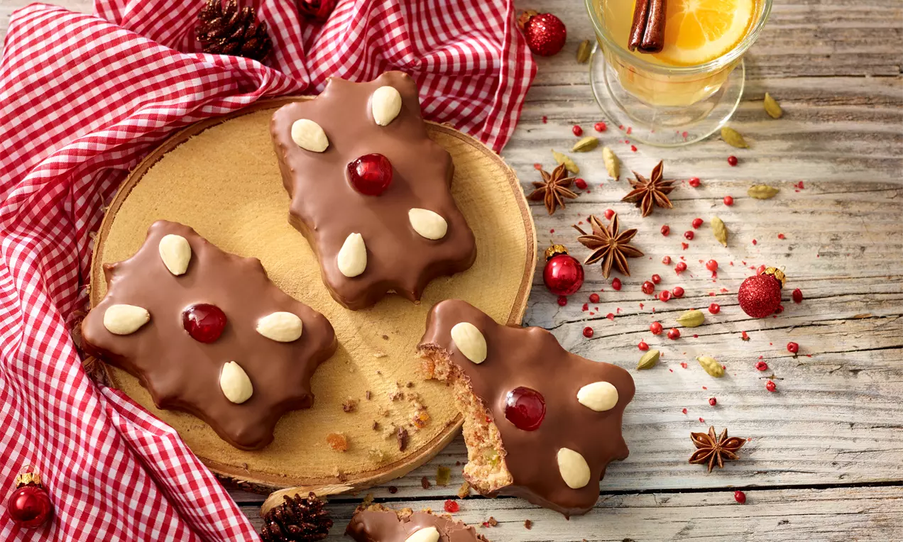 Lebkuchen mit dunkler Schokoladenglasur, ganzen Mandeln und roter kandierter Kirsche auf hellem Holzbrett mit rot-weiß kariertem Stoff drapiert