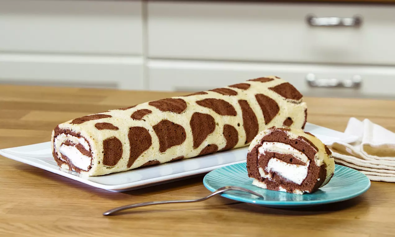 Roulade mit braunem Giraffenmuster und heller Füllung auf weißer Servierplatte, angeschnittener Teil auf türkisfarbenem Teller mit Gabel