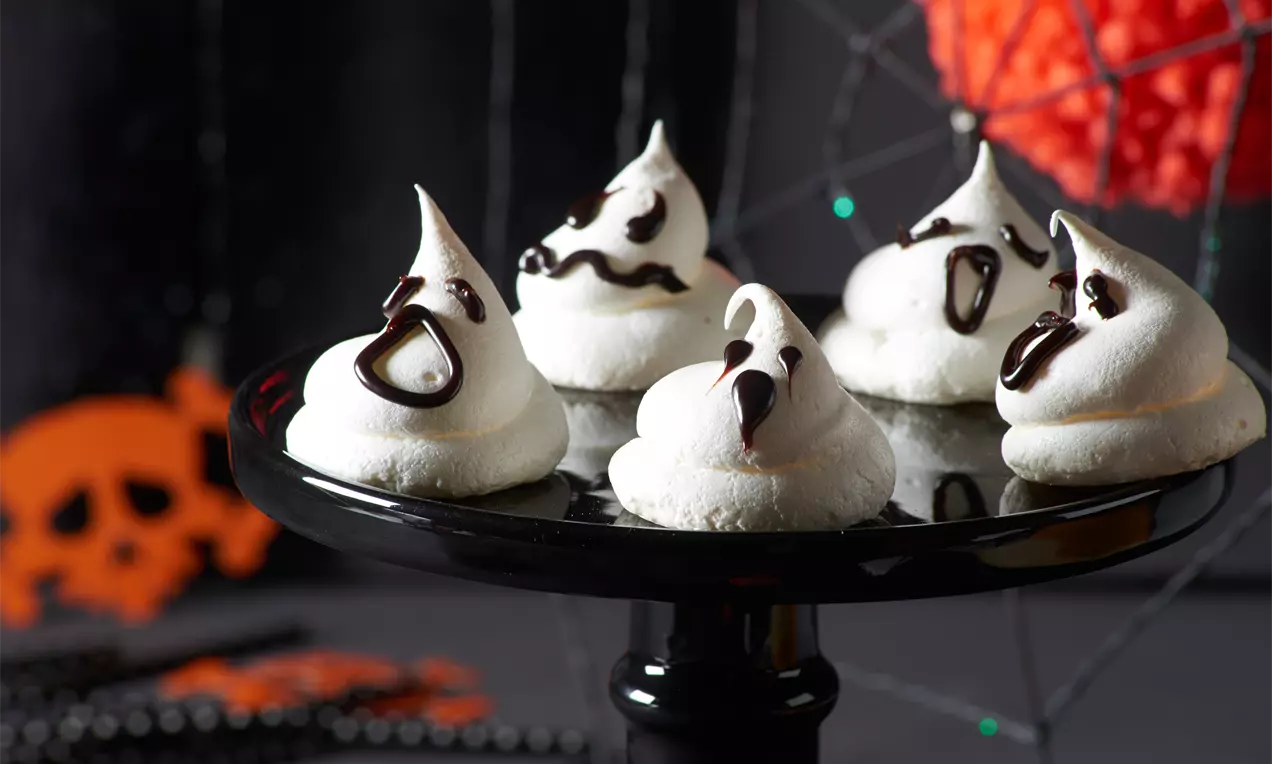 Weiße, spiralförmig gespritzte Baisers mit dunklen Schokoladenakzenten als Gesichter auf schwarzer Tortenplatte vor Halloween-Kulisse