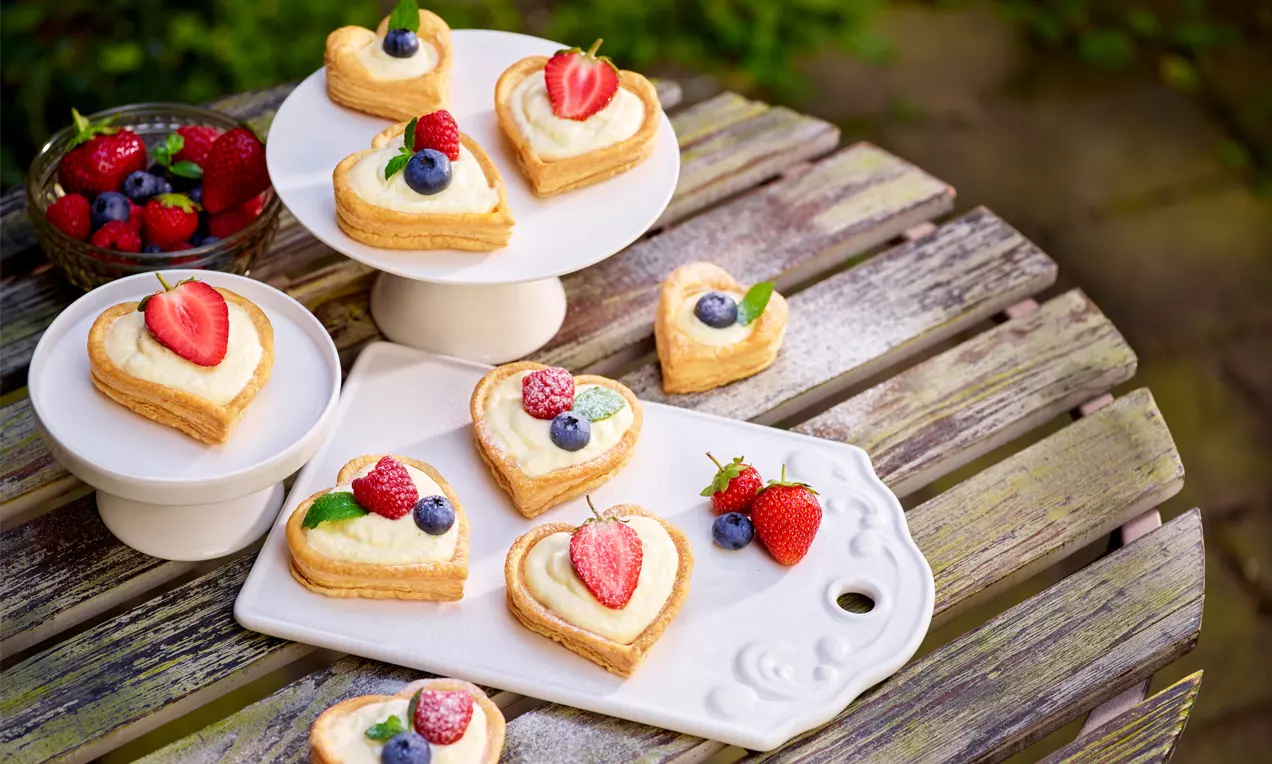 Goldene Blätterteig-Herzen mit flüssiger Cremefüllung dekoriert mit frischen Beeren auf weißen Servierplatten und rustikalem Holztisch