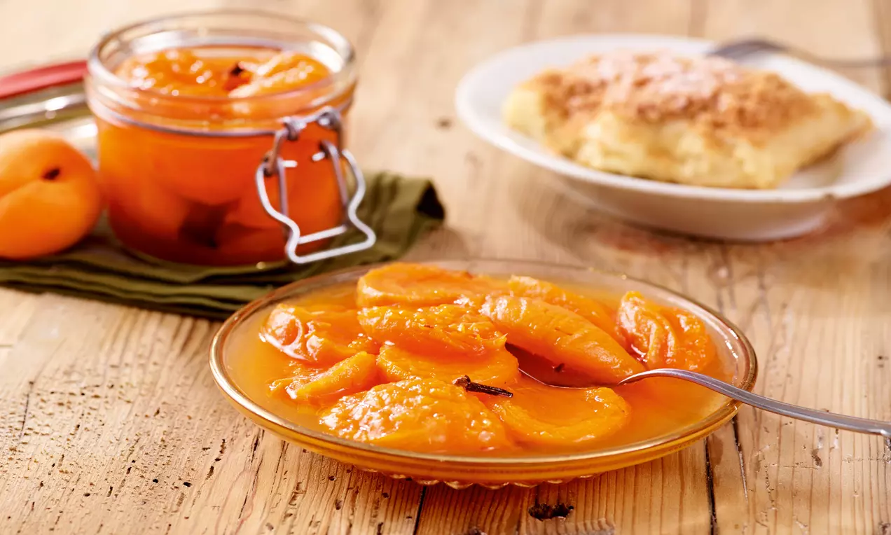 Marillenröster mit leuchtend orangefarbenen, weichen Fruchtstücken in sirupartigem Sud auf einem Glasteller, daneben ein Schraubglas und Kaiserschmarrn
