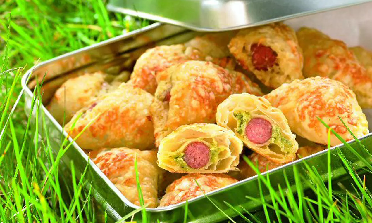 Goldbraune, zartblättrige Häppchen mit knuspriger Textur, gefüllt mit Wurst und grünem Pesto in einer silbernen Dose auf grünem Gras