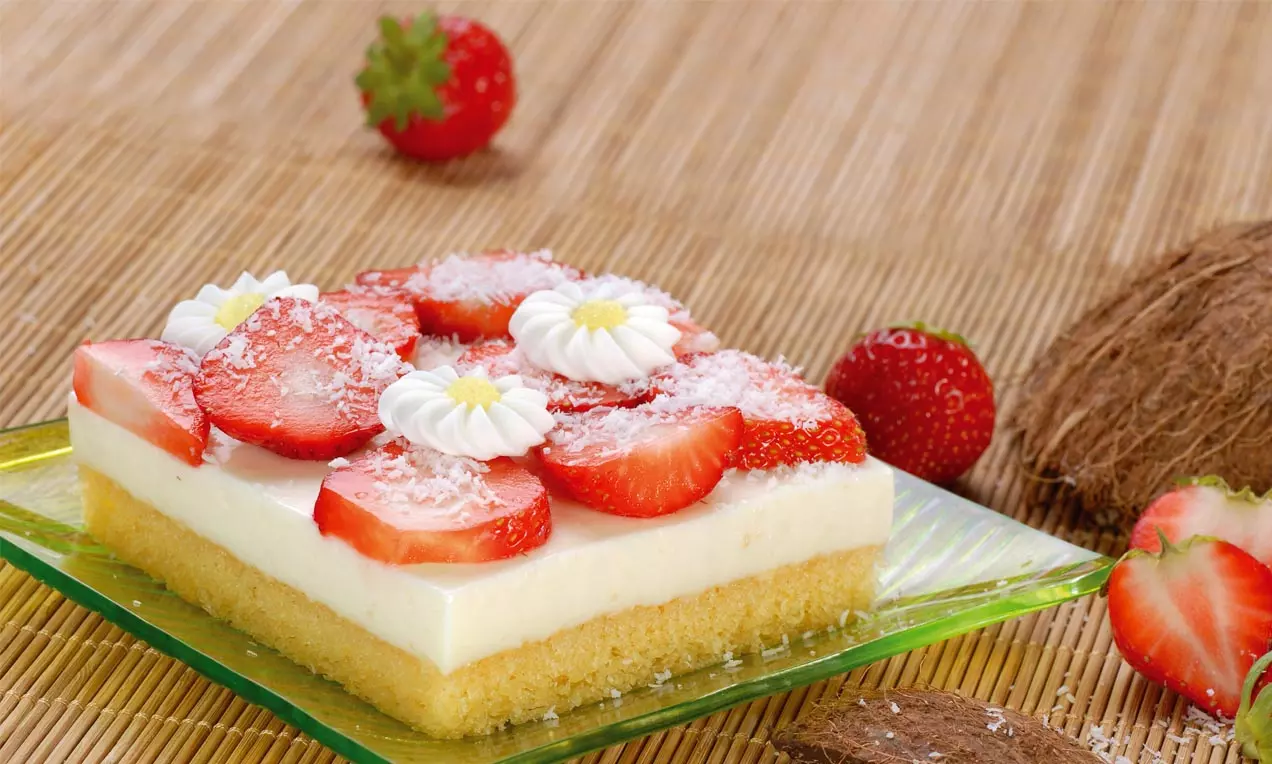 Rechteckiger Kokoskuchen mit heller Creme, frischen Erdbeerscheiben, weißen Blütendekoren und Kokosraspeln auf grünem Glasplatte