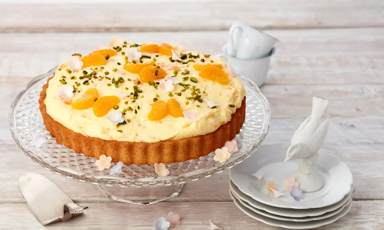 Schüttelkuchen mit goldbraunem Boden, heller Creme, Mandarinenstücken, gehackten Pistazien und Zuckerblüten auf gläserner Tortenplatte