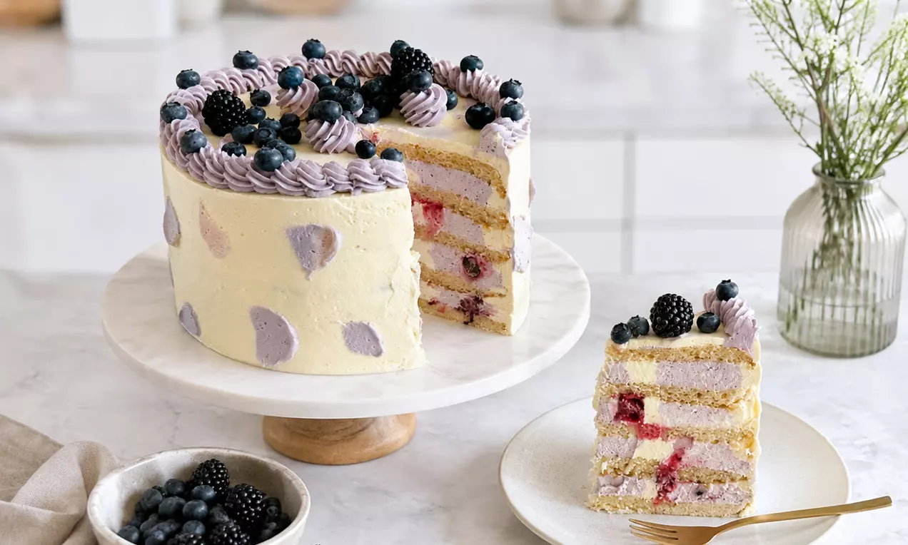 Helle vegane Buttercreme-Torte mit lilafarbenen Tupfen und Beeren dekoriert auf einer Tortenplatte, ein Stück auf einem Teller daneben, AI-labeled image