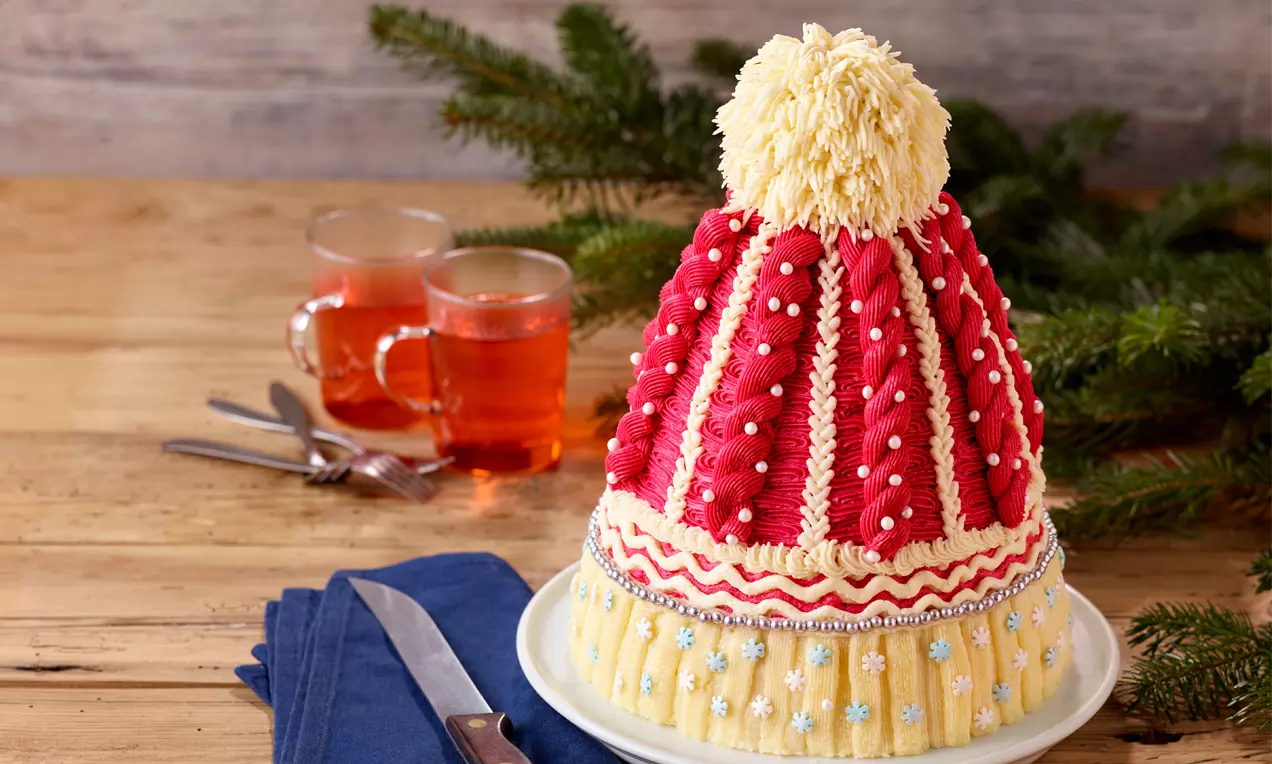 Kuppelförmiger Kuchen in Strickmützenoptik mit rotem und gelbem Frosting, weißen Perlen und gelber Haube auf weißem Teller mit Tannenzweigen