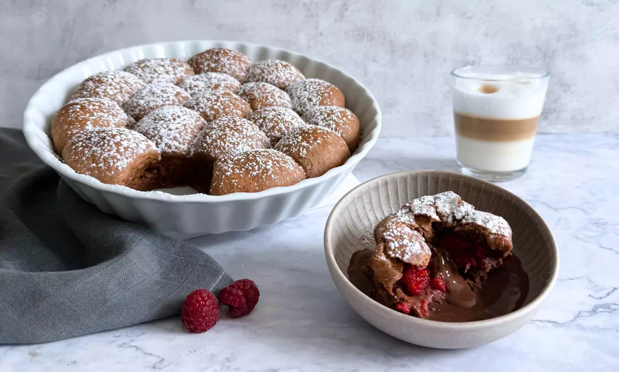 Fluffige, goldbraun gebackene Schoko-Buchteln mit Puderzucker bestäubt, angerichtet in einer weißen Schale, dazu Himbeeren und Latte Macchiato