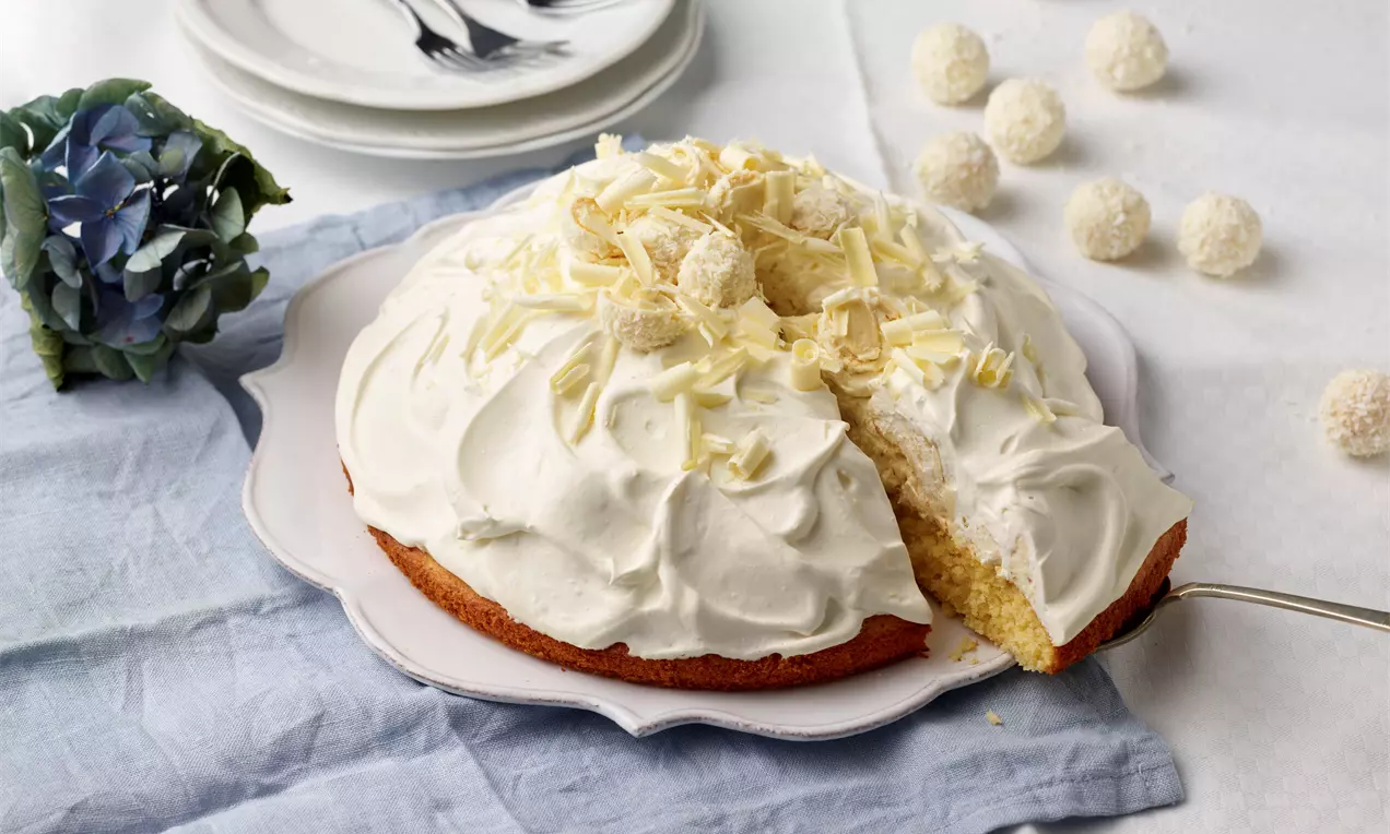 Raffaello-Torte mit weißer Sahnehaube, dekoriert mit geraspelter weißer Schokolade und Kugeln, auf einem Teller mit goldener Serviergabel
