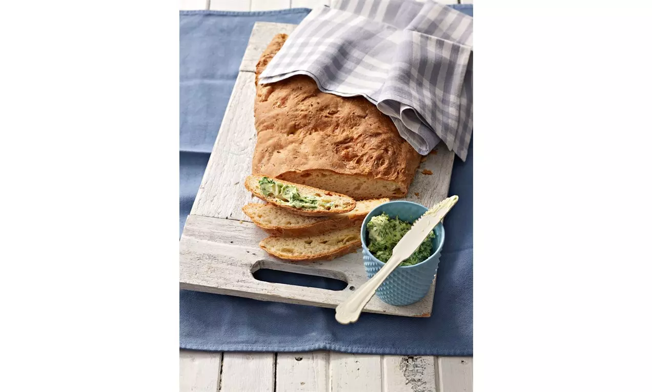 Drei-Käse-Brot mit goldbrauner Kruste, zwei angeschnittene Scheiben mit grüner Bärlauchbutter auf einem weißen Holzbrett mit blauem Tuch
