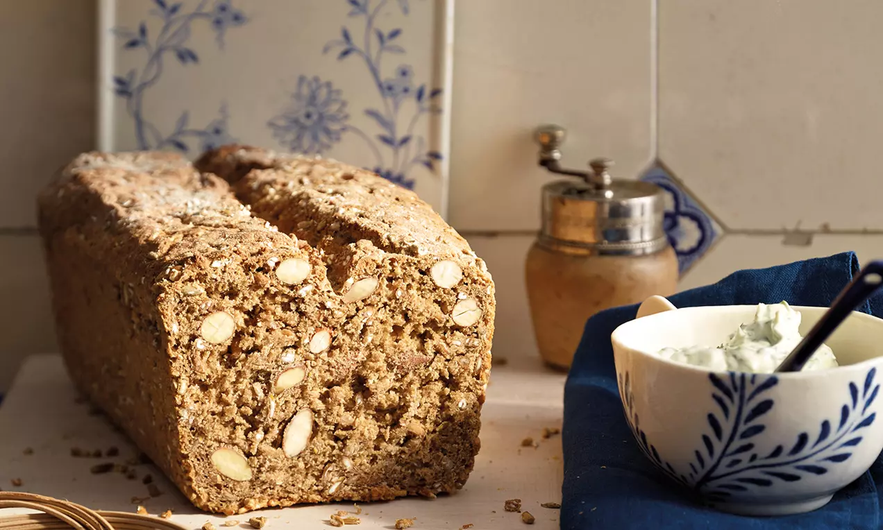 Roggenvollkornbrot mit körniger Textur und sichtbaren Saaten sowie Mandeln, auf einem weißen Untergrund mit blauem Geschirr drapiert