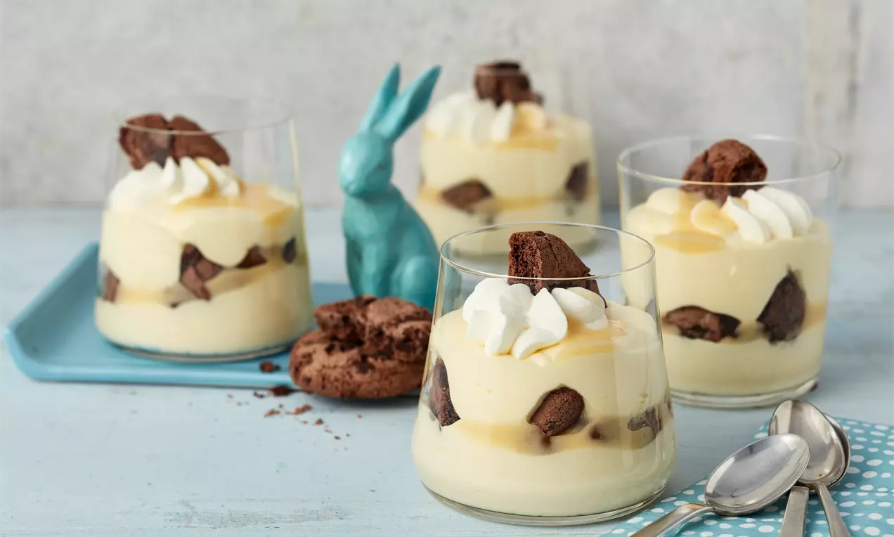 Gläser mit cremig gelber Eierlikörcreme, stückigem Schokoladenkuchen, Sahnetupfen und goldgelbem Eierlikör auf hellem Holz und blauer Deko