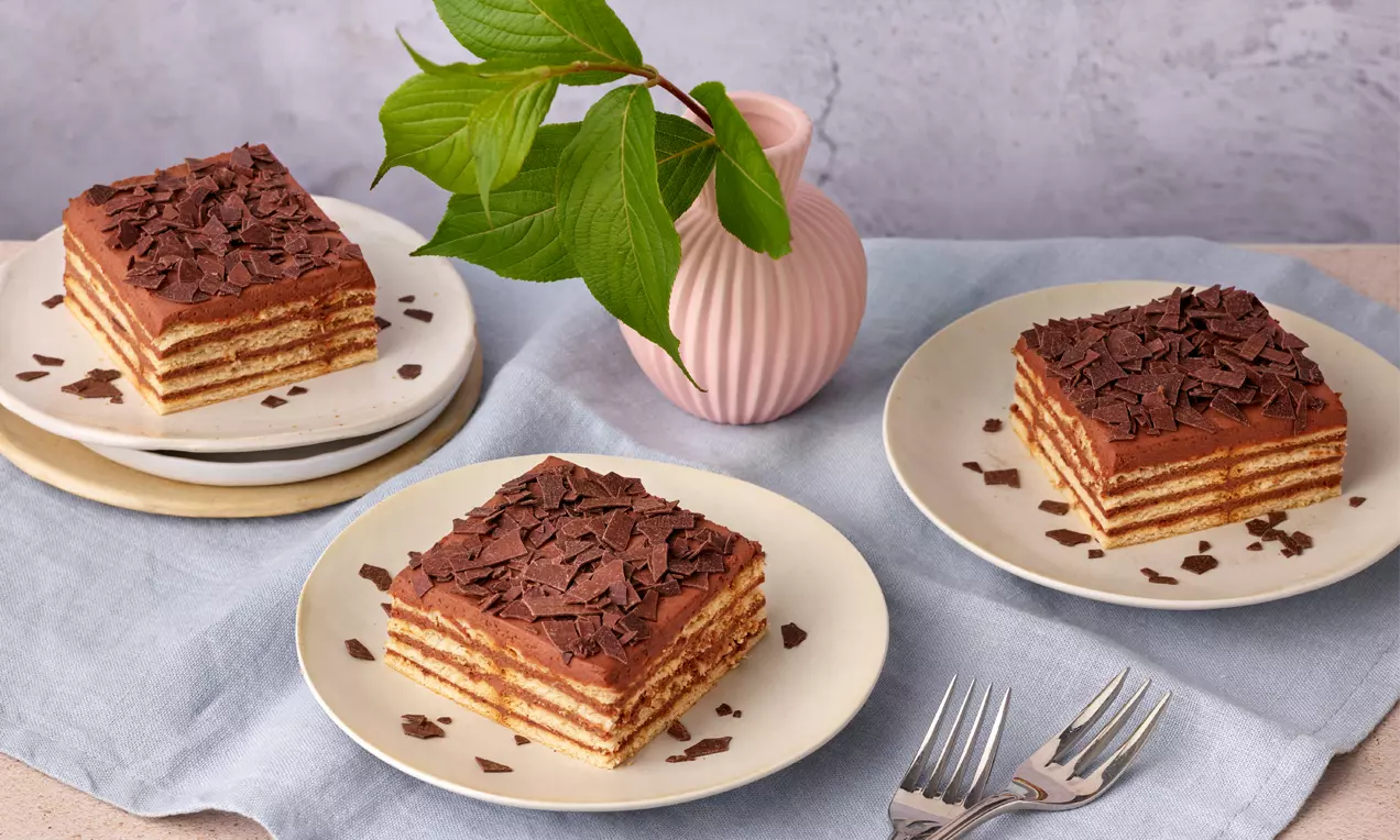 Quadratische Butterkeks-Schichten mit hellbrauner Creme und dunkler Schokoraspeldecke auf weißen Tellern, angerichtet mit blauem Stoff und rosa Vase