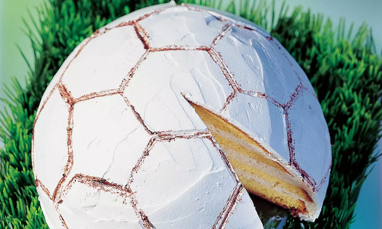 Kuchen in Form eines weißen Fußballs mit braunen Linien, angeschnitten, zeigt hellgelbe Schichten und liegt auf grünem Rasendekor