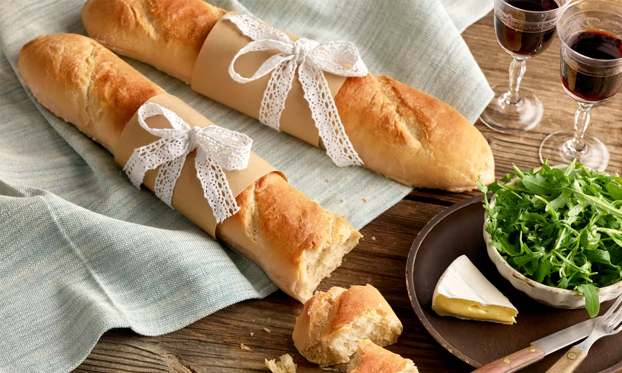 Zwei goldbraune Baguettes mit knuspriger Kruste, in Spitzenband gewickelt auf Leinentuch, neben Rucola, Brie und Rotweingläsern angerichtet