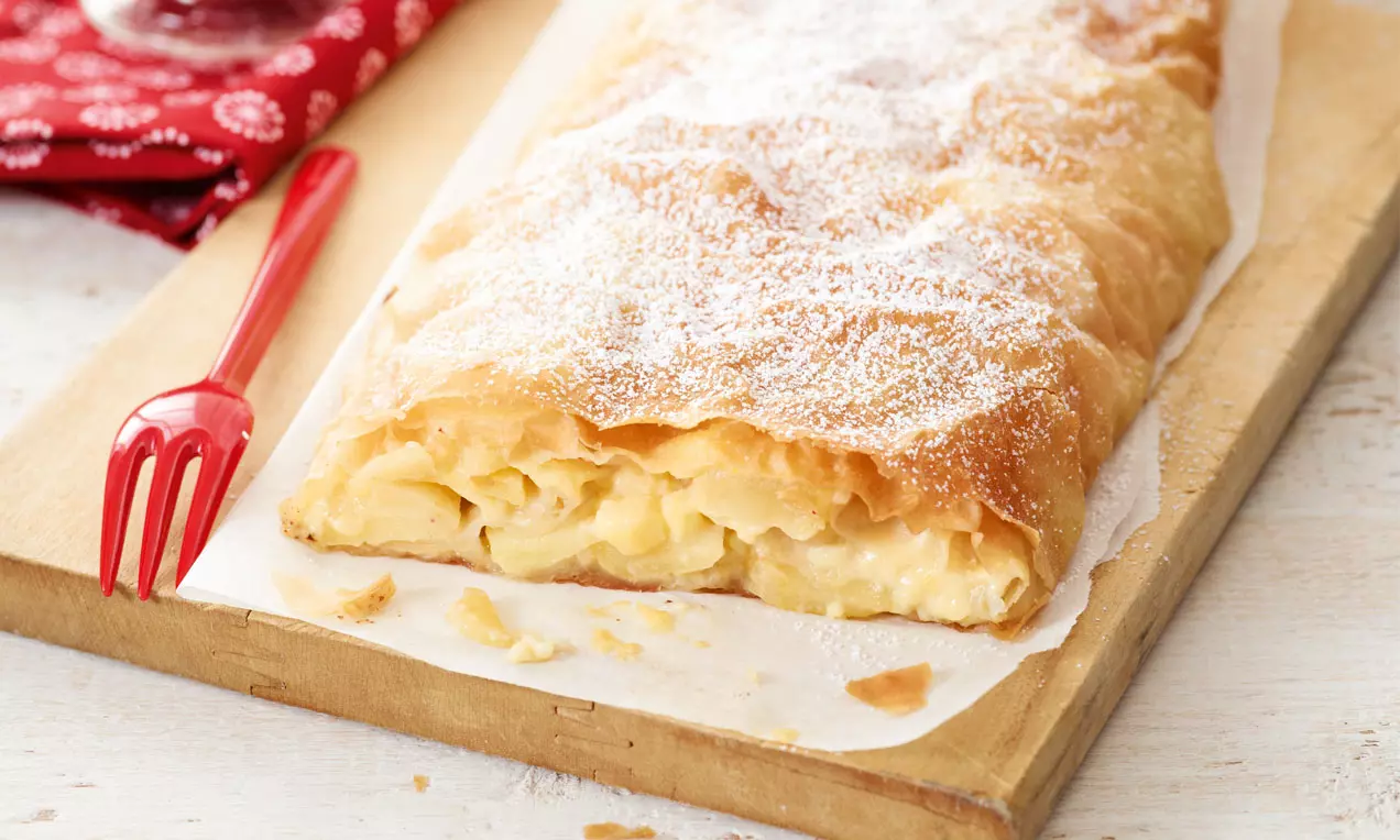 Goldbrauner Strudel mit zarter Apfel-Vanillepudding-Füllung, bestäubt mit Puderzucker, serviert auf hellem Holzbrett mit rotem Kuchengabel