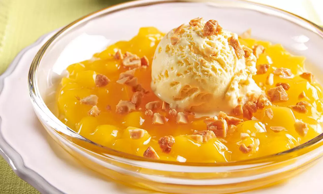 Vanilleeis-Kugel mit feiner Cremetextur und knusprigem Krokant auf leuchtend gelben, glänzenden Mangostückchen in einer Glasteller-Schale