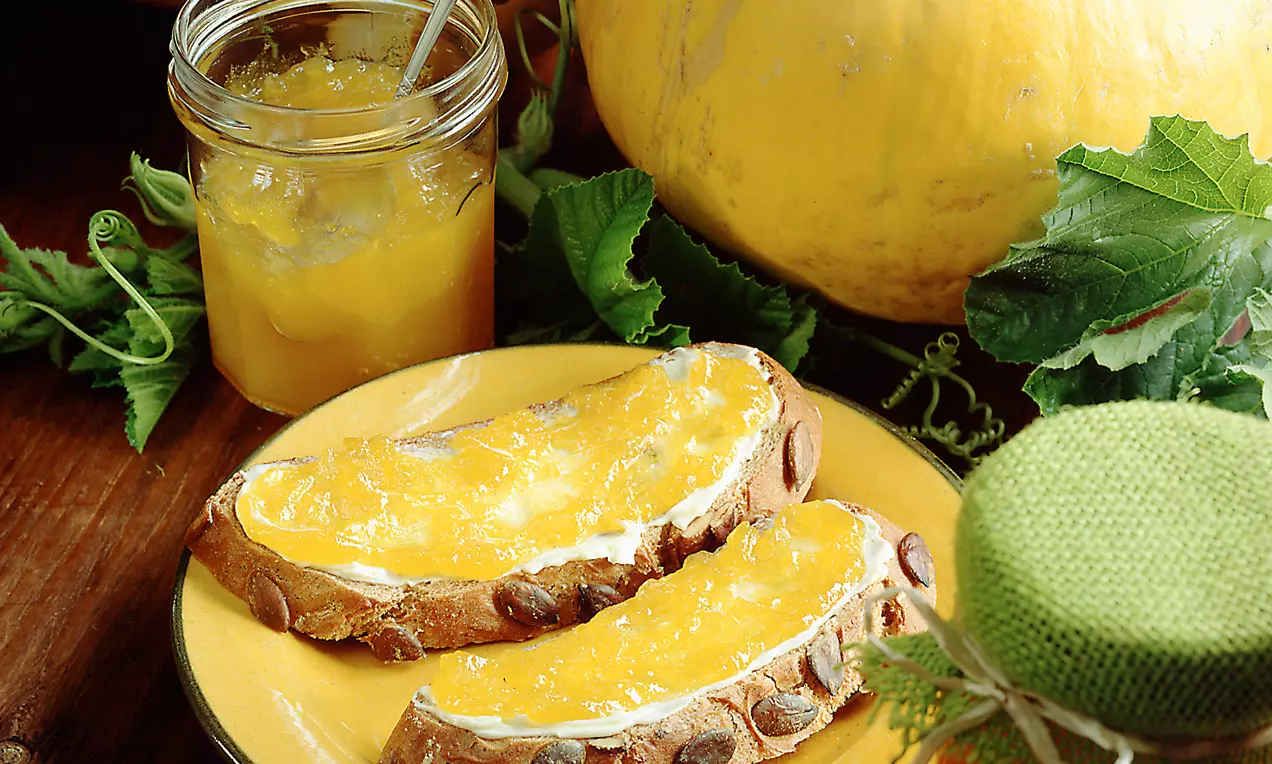 Ein Glas mit leuchtend gelber Kürbismarmelade neben einem Teller mit rustikalem Brot, bestrichen mit der Marmelade, umgeben von Kürbis und grünen Blättern