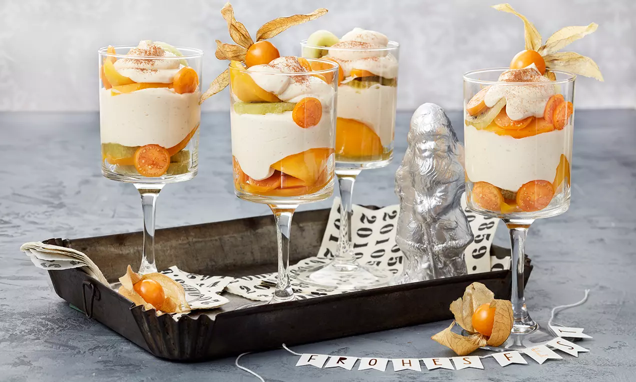 Schichtdessert in Gläsern mit heller Zimt-Orangencreme, frischen Früchten wie Physalis und Melone, dekoriert mit Zimt und Physalis-Blättern