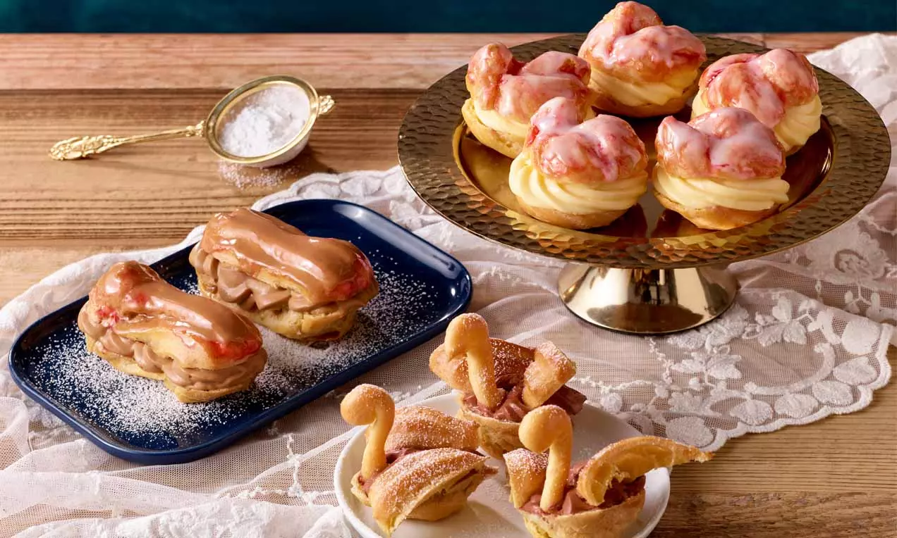 Brandteigkrapfen mit rosa Glasur auf goldener Etagere, helle Eclairs mit karamellfarbener Creme und gezuckerte Schwäne auf Spitzenstoff drapiert