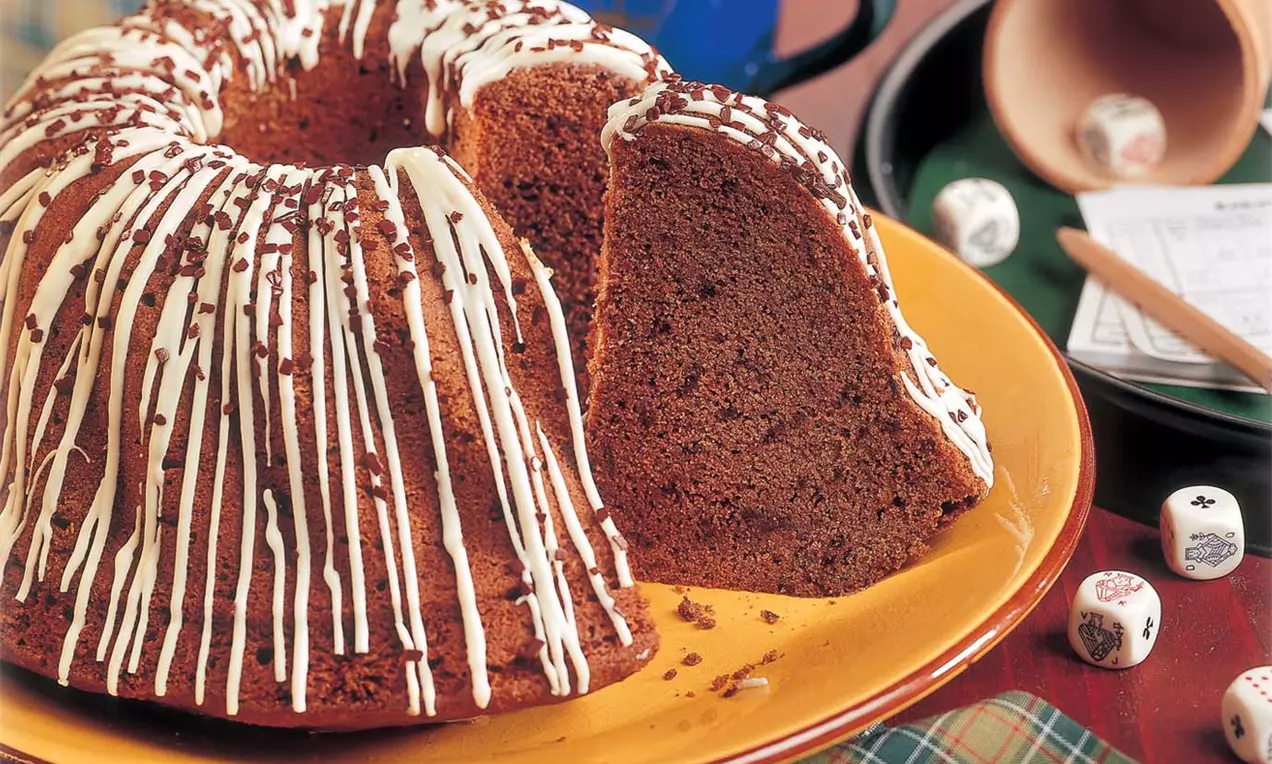 Dunkelbrauner Gugelhupf mit heller Zuckerglasur in Streifen, eine große Scheibe herausgeschnitten, auf gelbem Teller mit Spielwürfeln drapiert