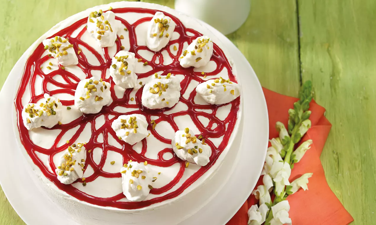 Holunderblüten-Sektcremetorte mit weißer Creme, roten Fruchtsoßenlinien, Sahnetupfen und gehackten Pistazien auf weißer Platte