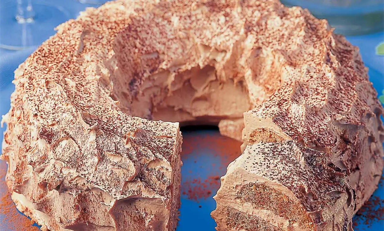 Runder Schichtkuchen mit cremiger hellbrauner Oberfläche und Kakaopulver bestäubt, auf blauem Tuch mit Kleeblättern und Kerze verziert