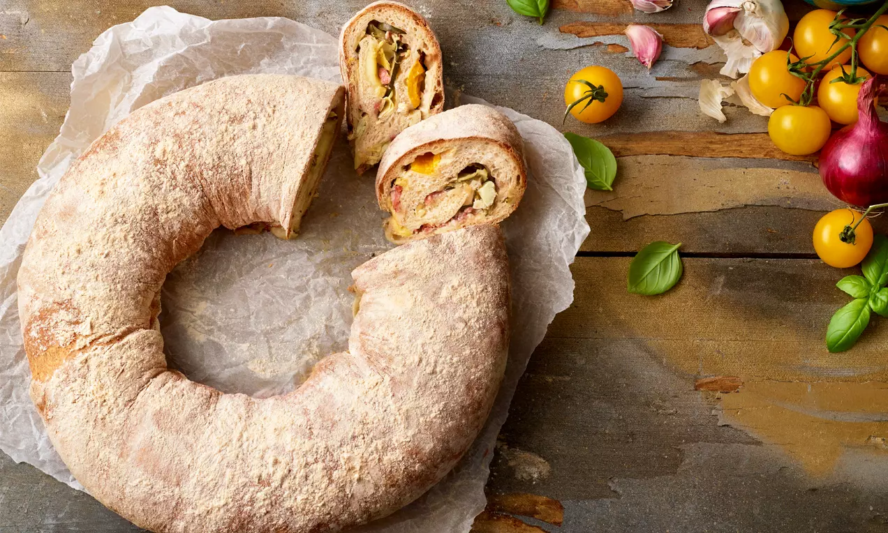 Rustikaler goldbrauner Brotkranz mit mehliger Kruste, gefüllt mit buntem Gemüse, auf Backpapier und Holztisch neben frischen Tomaten und Kräutern
