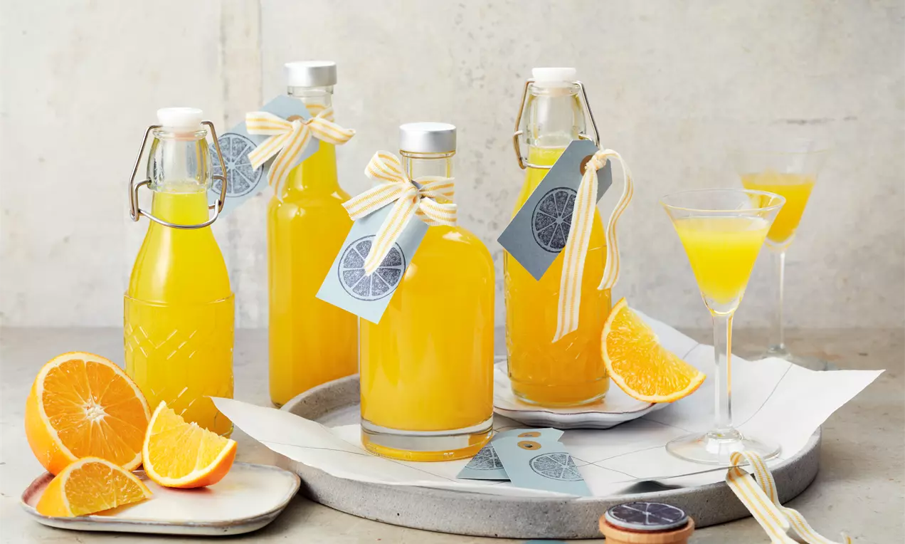 Leuchtend orangefarbener Likör in Glasflaschen mit weiß-gelber Schleife und Etikett, arrangiert mit frischen Orangenscheiben auf hellem Untergrund