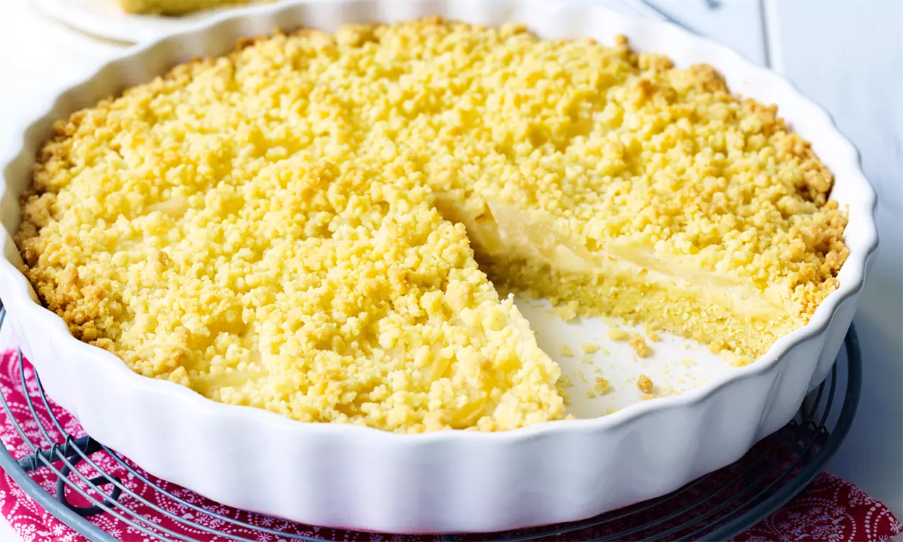 Goldgelbe Birnen-Marzipan-Tarte mit buttrigen Streuseln, in einer weißen Tarteform auf einem roten Stoff mit feinem Muster serviert