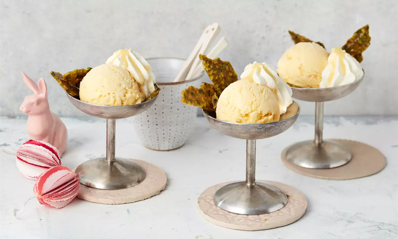 Drei silberne Dessertschalen mit gelbem Eierliköreis, Sahnehäubchen und knusprigen Krokantstücken vor hellem Hintergrund arrangiert