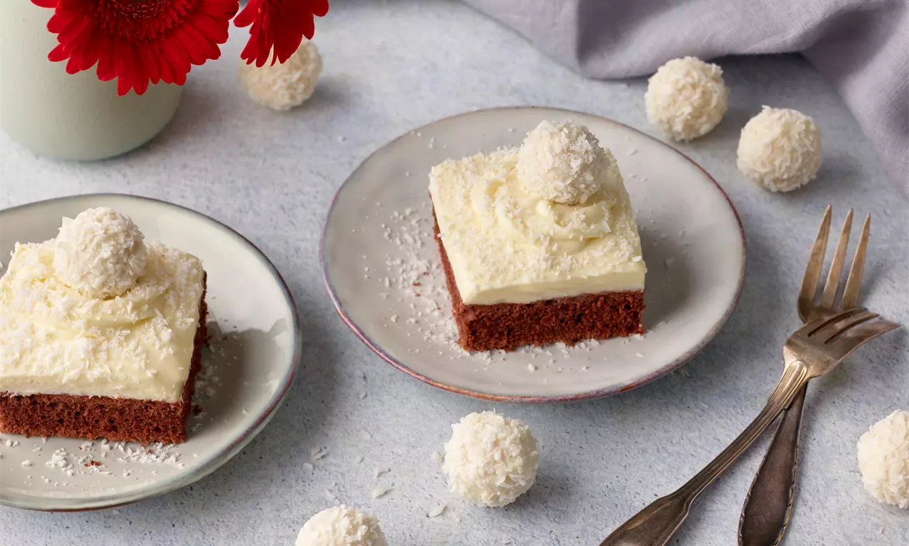 Saftige, hellbraune Kuchenquadrate mit cremiger, weißer Schicht, Kokosraspeln und Raffaello-Kugeln auf Tellern, umgeben von roten Blumen