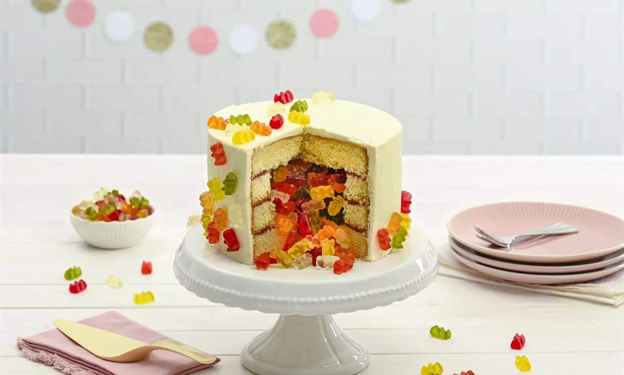 Weiße Schichttorte mit hellem Biskuit und farbigen Goldbären im Inneren, garniert mit Goldbären auf einer weißen Tortenplatte