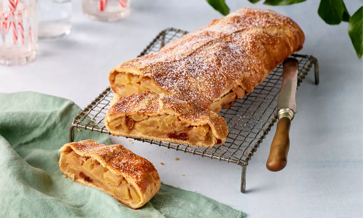 Goldbrauner Apfelstrudel aus Mürbteig mit Faltenstruktur und Puderzucker bestäubt, auf Gitter mit grünem Tuch und Messer angerichtet
