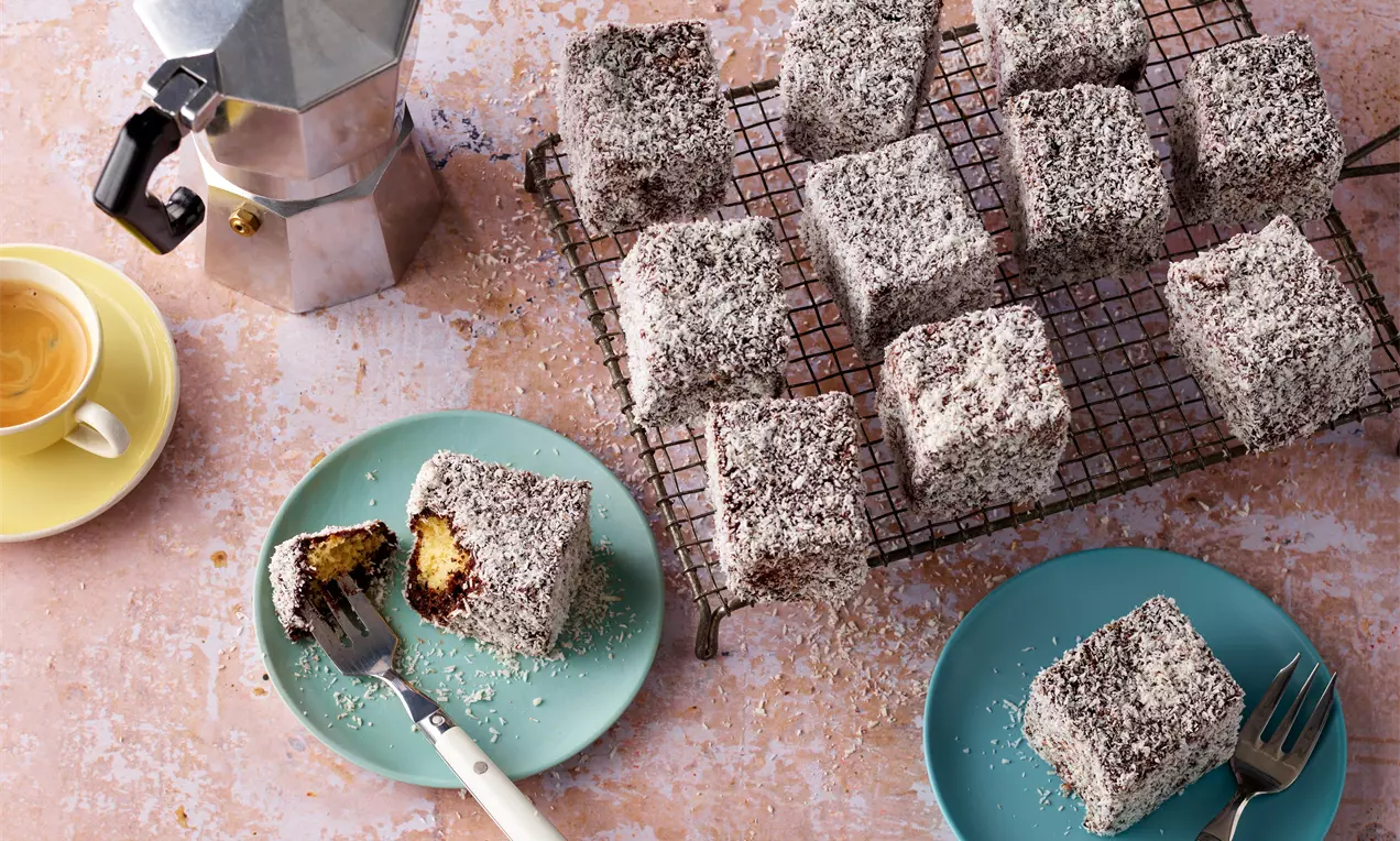 Würfelförmige Kuchen mit Schokoladenglasur und Kokosraspeln auf einem Gitter, daneben Kaffee und einzelne Stücke auf bunten Tellern