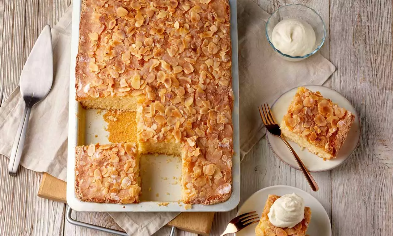 Goldbrauner Buttermilchkuchen mit glasierten Mandelblättchen in rechteckiger Form, angeschnitten und auf Tellern mit Sahne serviert