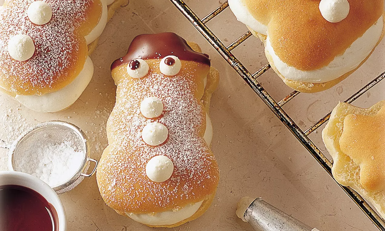 Schneemannförmige Gebäckteile mit golden gebackener Oberfläche, weißer Füllung, Schokoladehut, Augen und Puderzucker auf einem Gitter