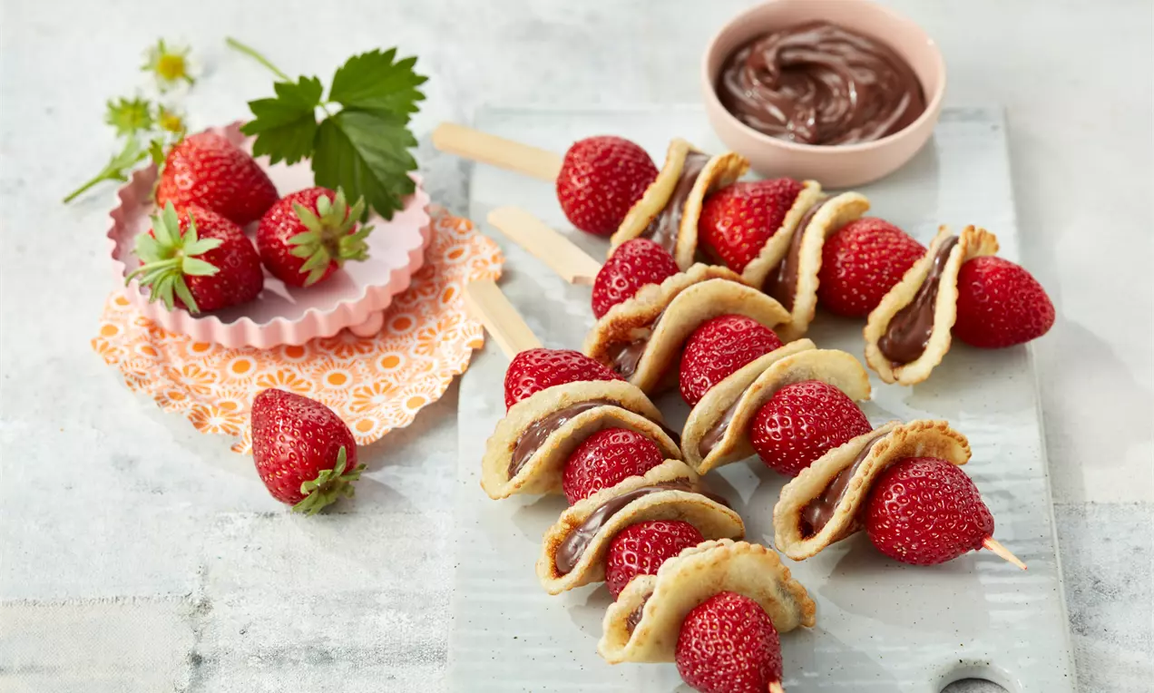 Auf Holzspießen gereihte rote Erdbeeren und goldbraune Püfferchen mit Schokoladencreme, arrangiert auf hellem Untergrund mit Schälchen und Dekoration