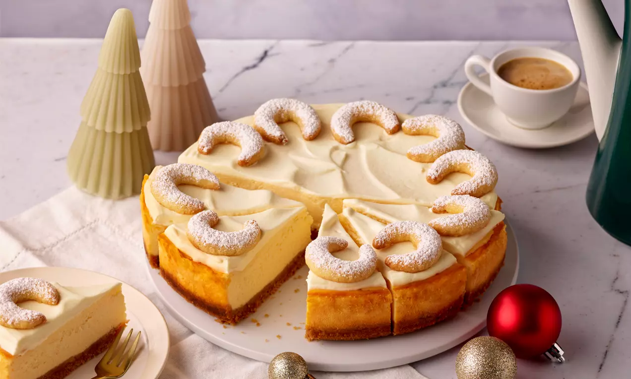 Vanillekipferl-Cheesecake mit cremig-heller Füllung auf knusprigem Boden, dekoriert mit Puderzucker-Kipferln auf weißer Tortenplatte