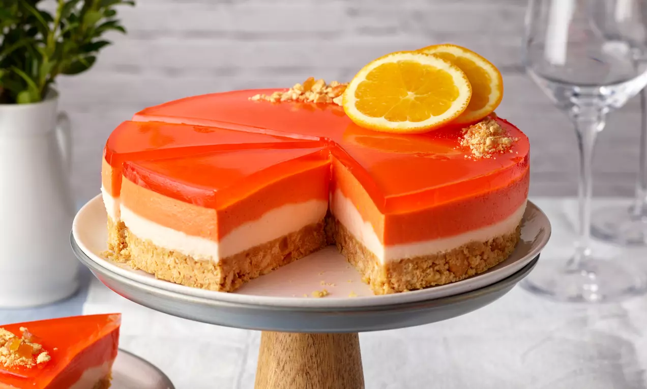 Aperol-Spritz-Frischkäsetorte mit orangefarbenem Gelee, heller Schichtfüllung und Keksboden auf einer weißen Tortenplatte mit Orangenscheiben garniert