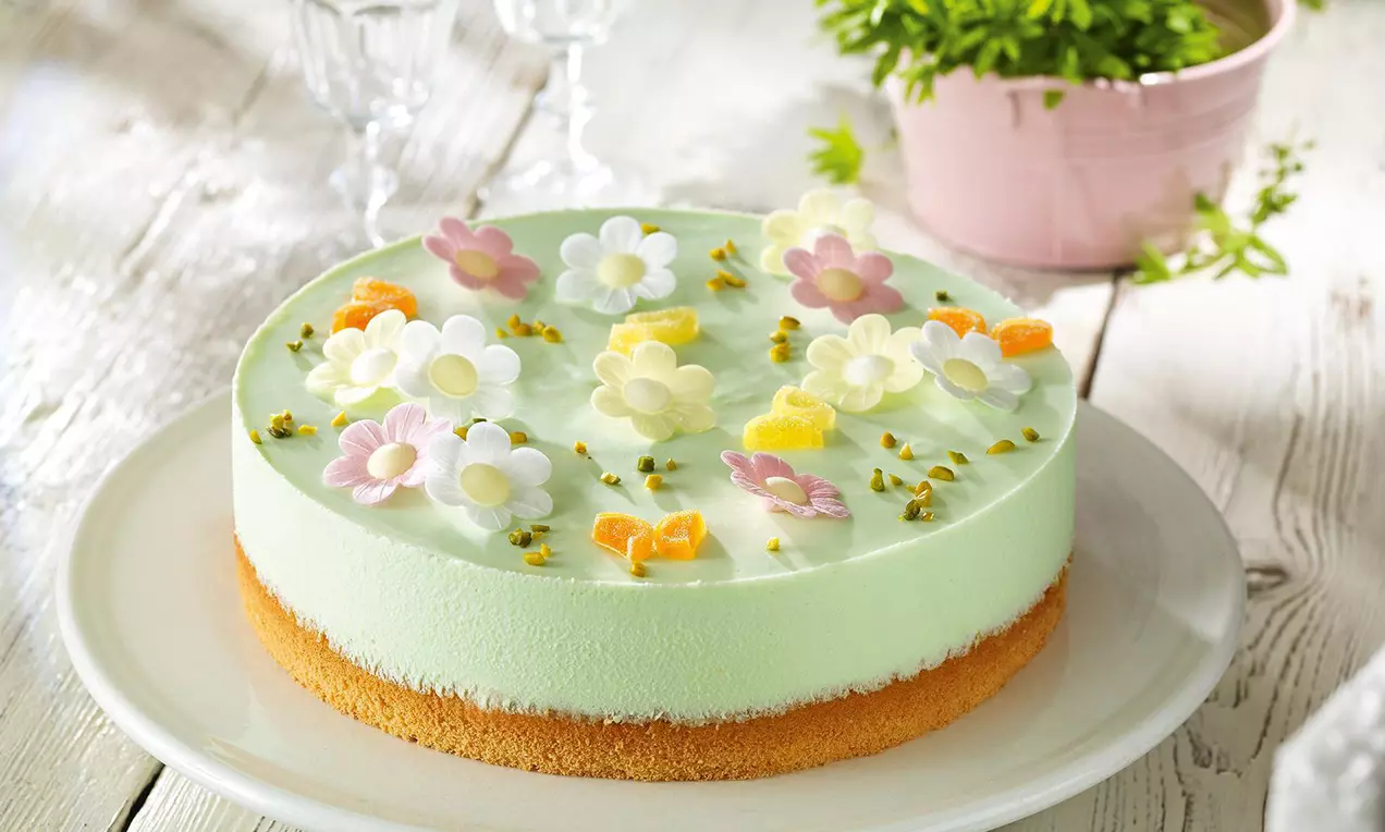 Rund geformte Torte mit grünlicher Creme und Blüten-Dekor aus Zucker, auf hellem Biskuitboden und weißem Teller angerichtet